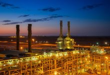 Vista aérea de uma instalação industrial iluminada ao entardecer, com edifícios e chaminés.